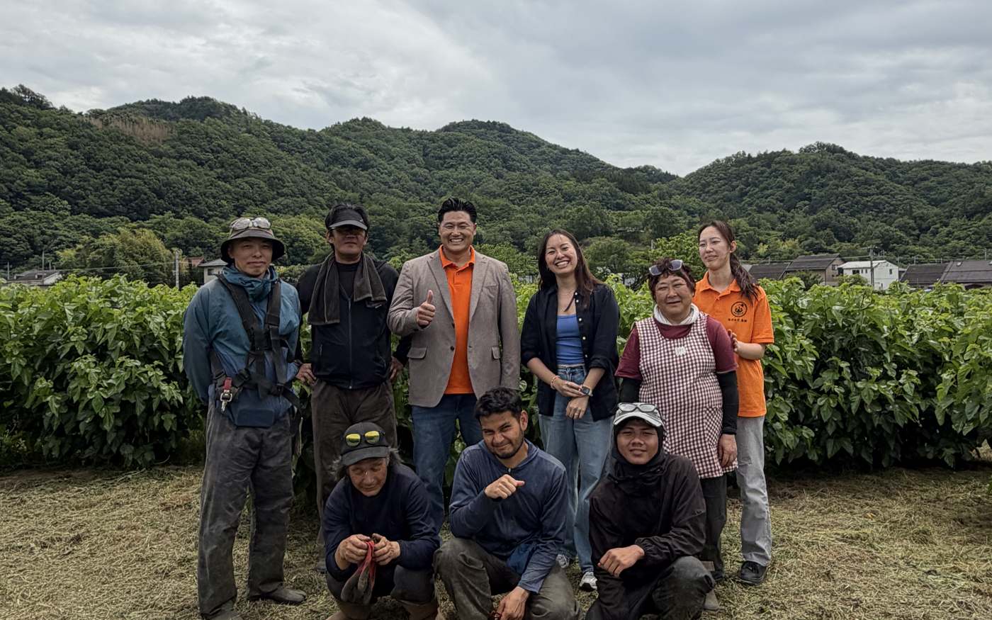 Keika and a farm team smiling for a group photo in front of mulberry rows, mountains behind