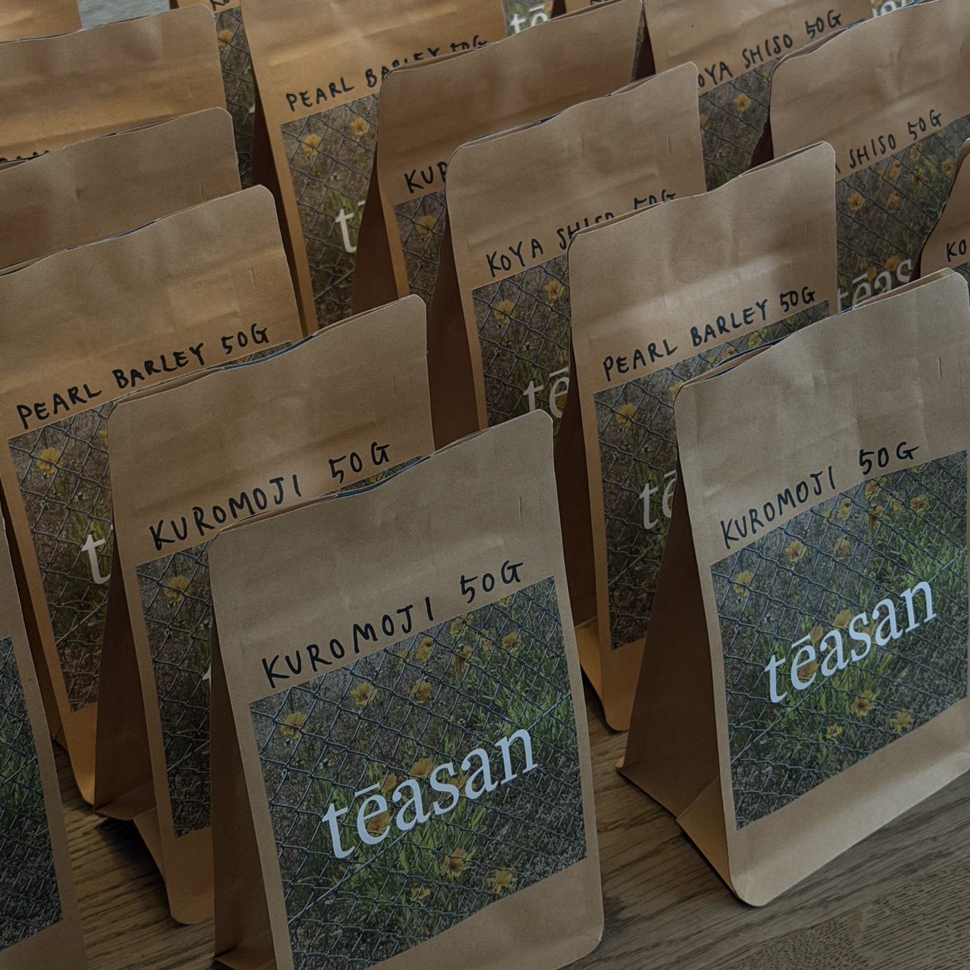 Kraft pouches of Kuromoji, Pearl Barley and Koya Shiso, hand-labelled 50g, lined up on a wooden table