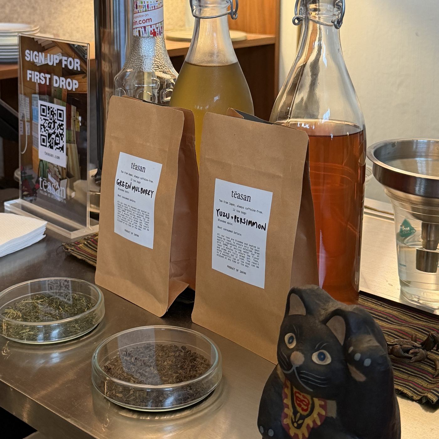 Two kraft paper Teasan tea pouches beside glass petri dishes of dry tea leaf and bottles of brewed tea